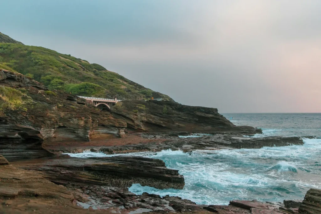 Waves crashing on layered rock shelves beneath a highway arch bridge on Oahu's coast
