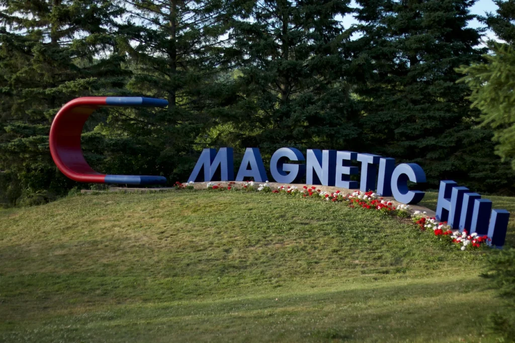A large outdoor sign reading Magnetic Hill next to a giant red and blue horseshoe magnet sculpture on a grassy hill