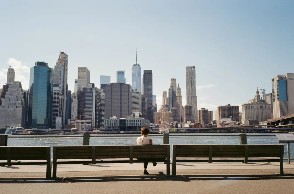 Person sitting on a wooden park bench looking at the Manhattan skyline across the river