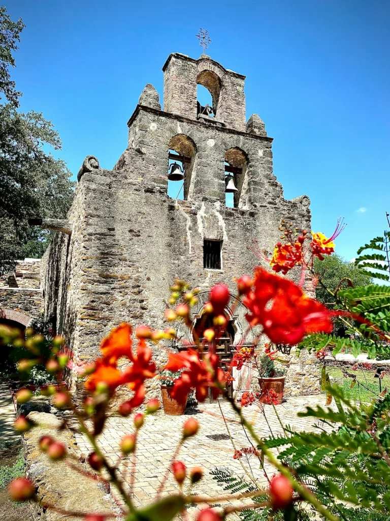 Historic stone church facade of Mission Espada framed by bright orange flowers under a clear blue sky