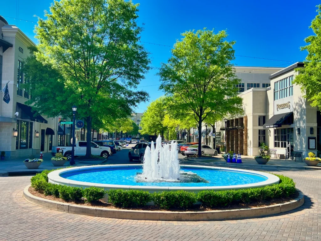 A bright blue circular fountain on a brick street lined with shops and green trees at North Hills in Raleigh