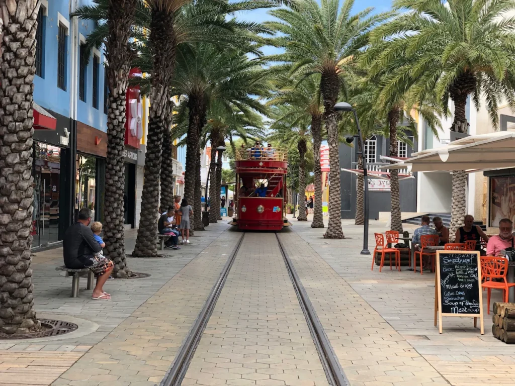 Red double-decker tram traveling down a brick pedestrian street lined with palm trees in Oranjestad