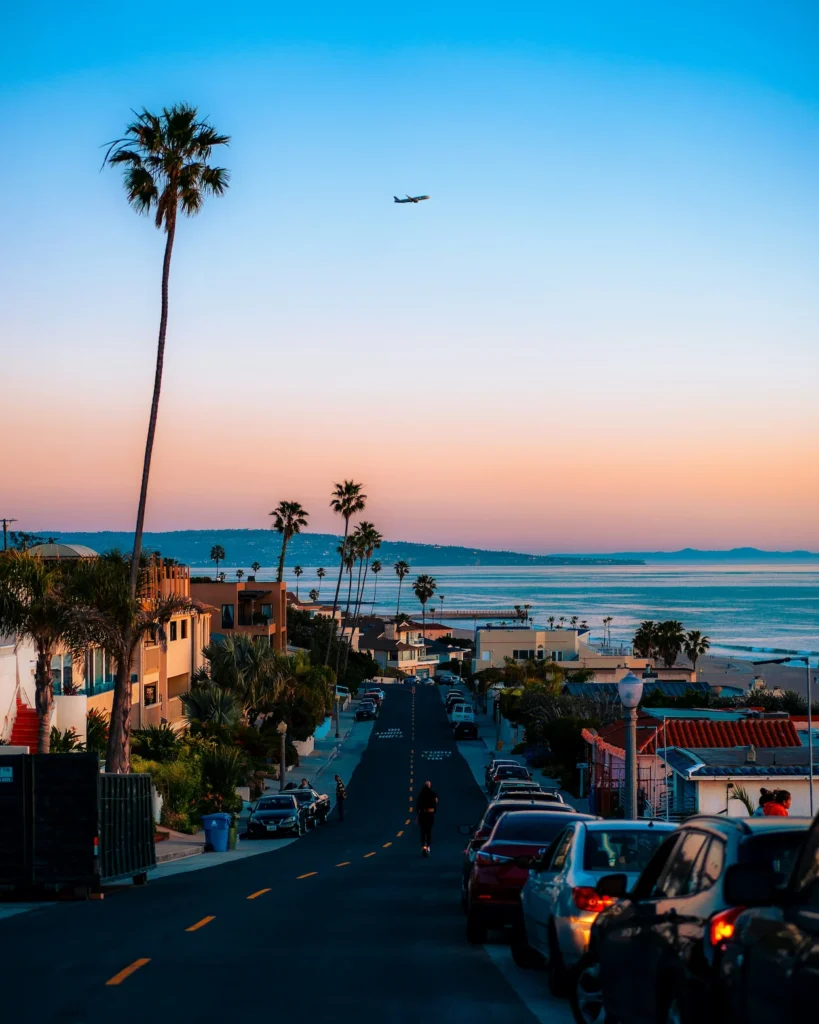 Coastal street lined with palm trees and parked cars looking down toward the ocean at sunset
