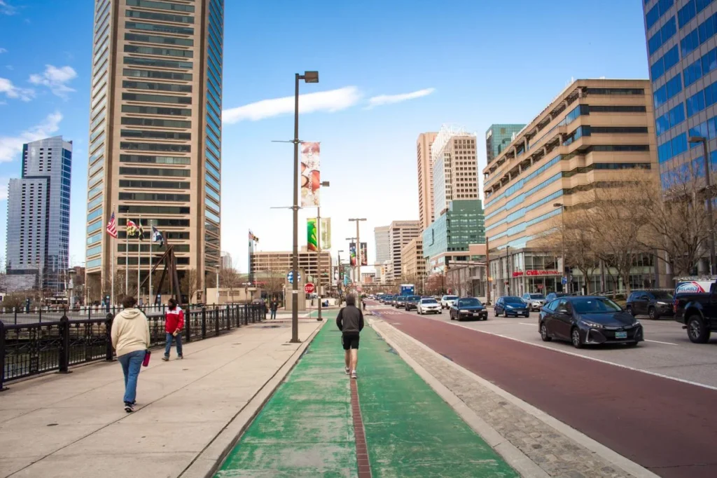 Pedestrians walking past skyscrapers on a wide sidewalk with a green bike lane in downtown Baltimore