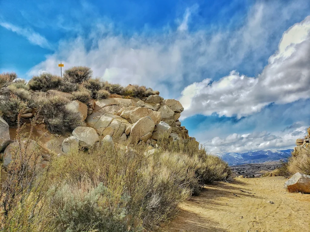A dirt path winding through desert sagebrush and large rock formations under a cloudy blue sky in Reno, Nevada