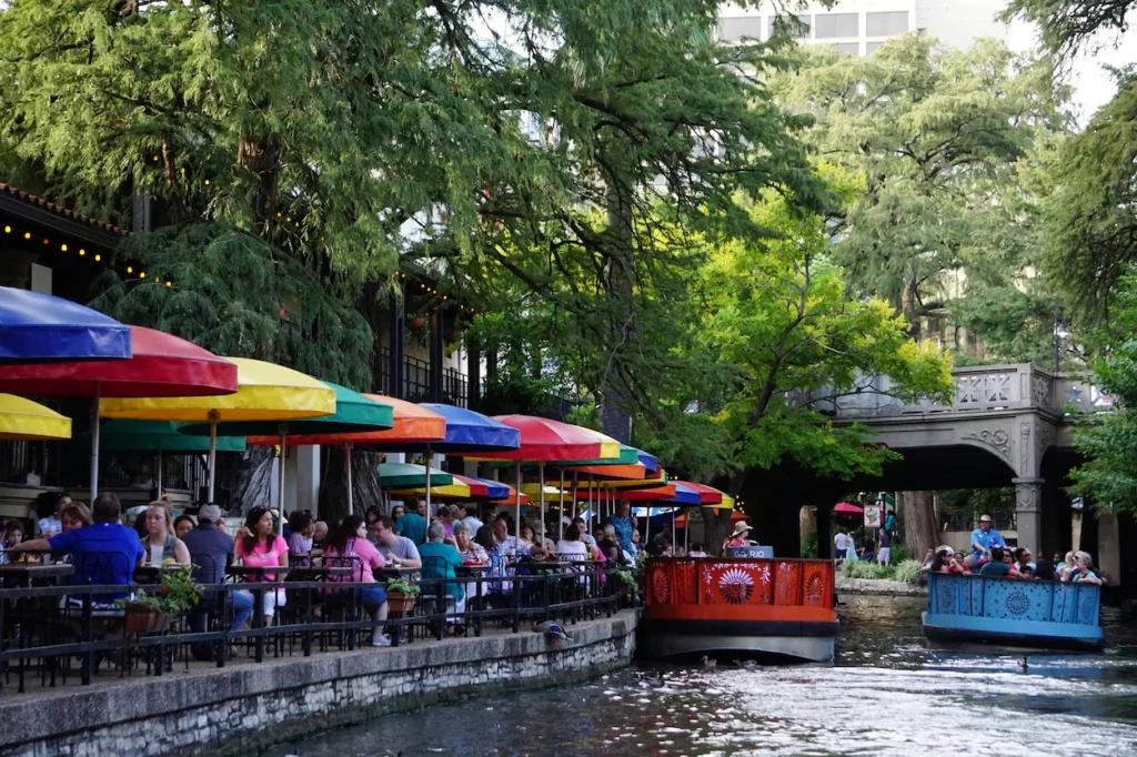 Tour boats passing riverside restaurants with colorful umbrellas along the San Antonio River Walk