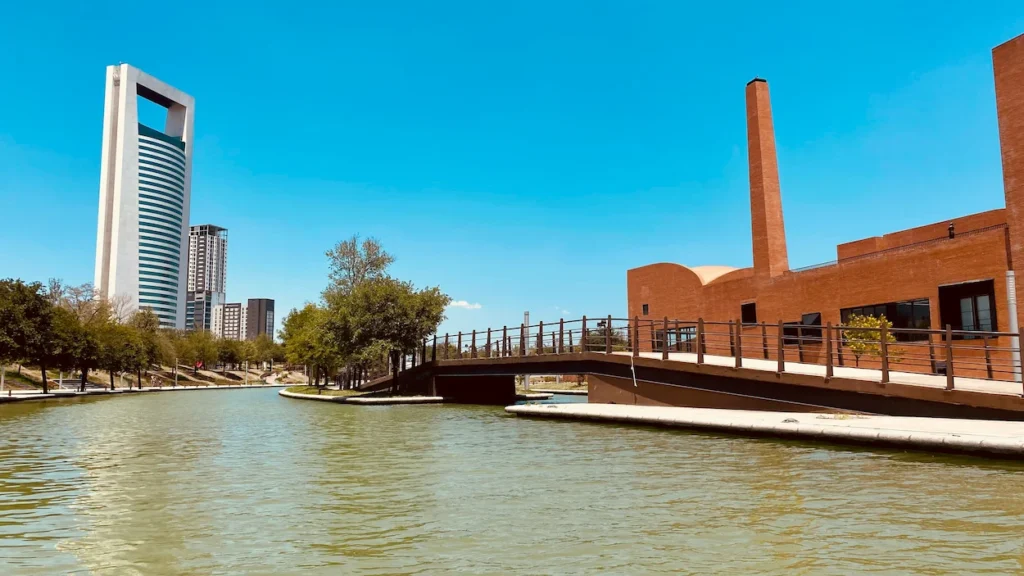 Man-made canal flowing past a brick building and a pedestrian bridge with modern city skyscrapers in the background