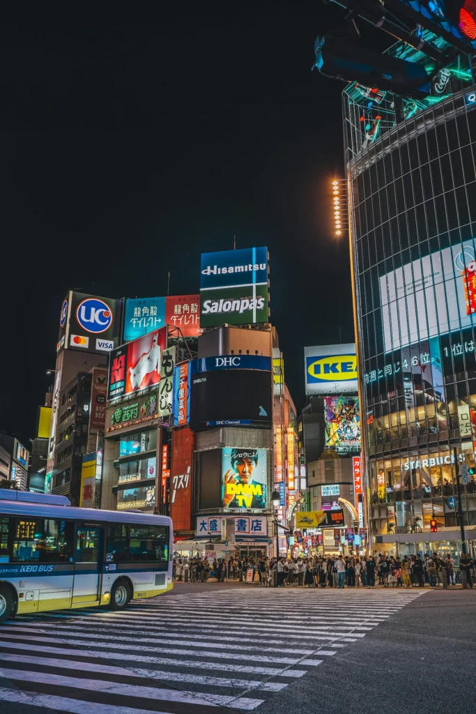 Shibuya Crossing intersection at night with glowing neon advertisements and a Keio Bus in the foreground