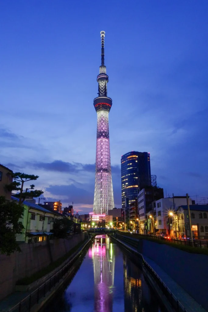 Illuminated Tokyo Skytree tower reflecting pink and white lights on a river canal at twilight