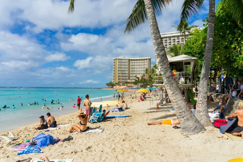 Crowded sandy shore of Waikiki Beach in Honolulu with palm trees, a lifeguard tower, and high-rise hotels