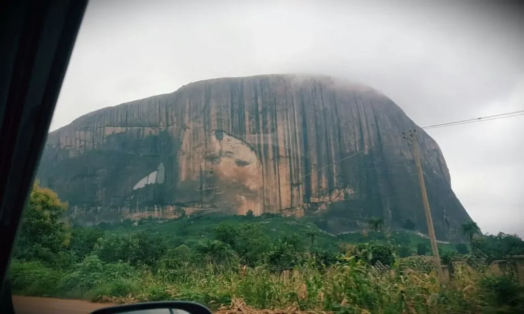 A view of the massive Zuma Rock with its distinctive face-like markings under a cloudy sky.