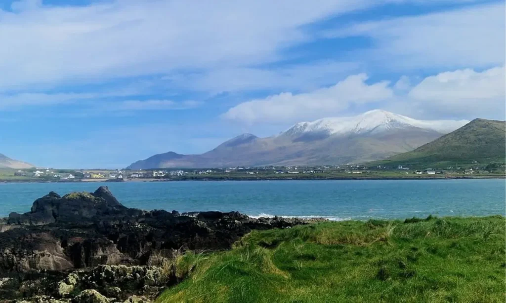 Dingle Peninsula shows a scenic coastline with grassy cliffs, blue water, and snow-capped mountains in the distance.