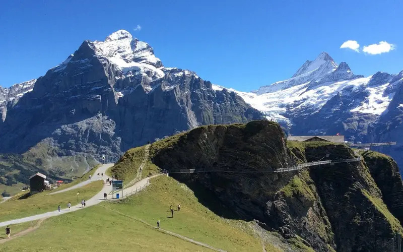 Hikers walk near First Mountain with snowy alpine peaks behind.