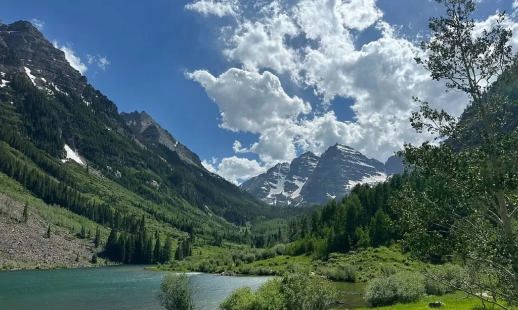 Snow-capped Maroon Bells mountains rise above a green valley and lake.