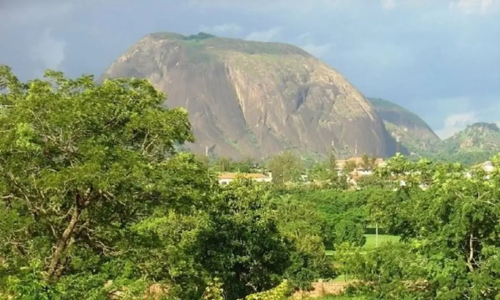 A scenic view of Abuja with lush greenery in the foreground and a large rocky hill in the background.