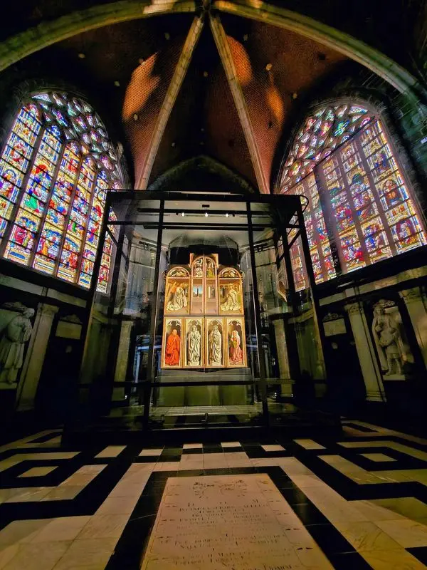 The Ghent Altarpiece at St. Bavo’s Cathedral displayed inside a glass case between two large stained-glass windows.
