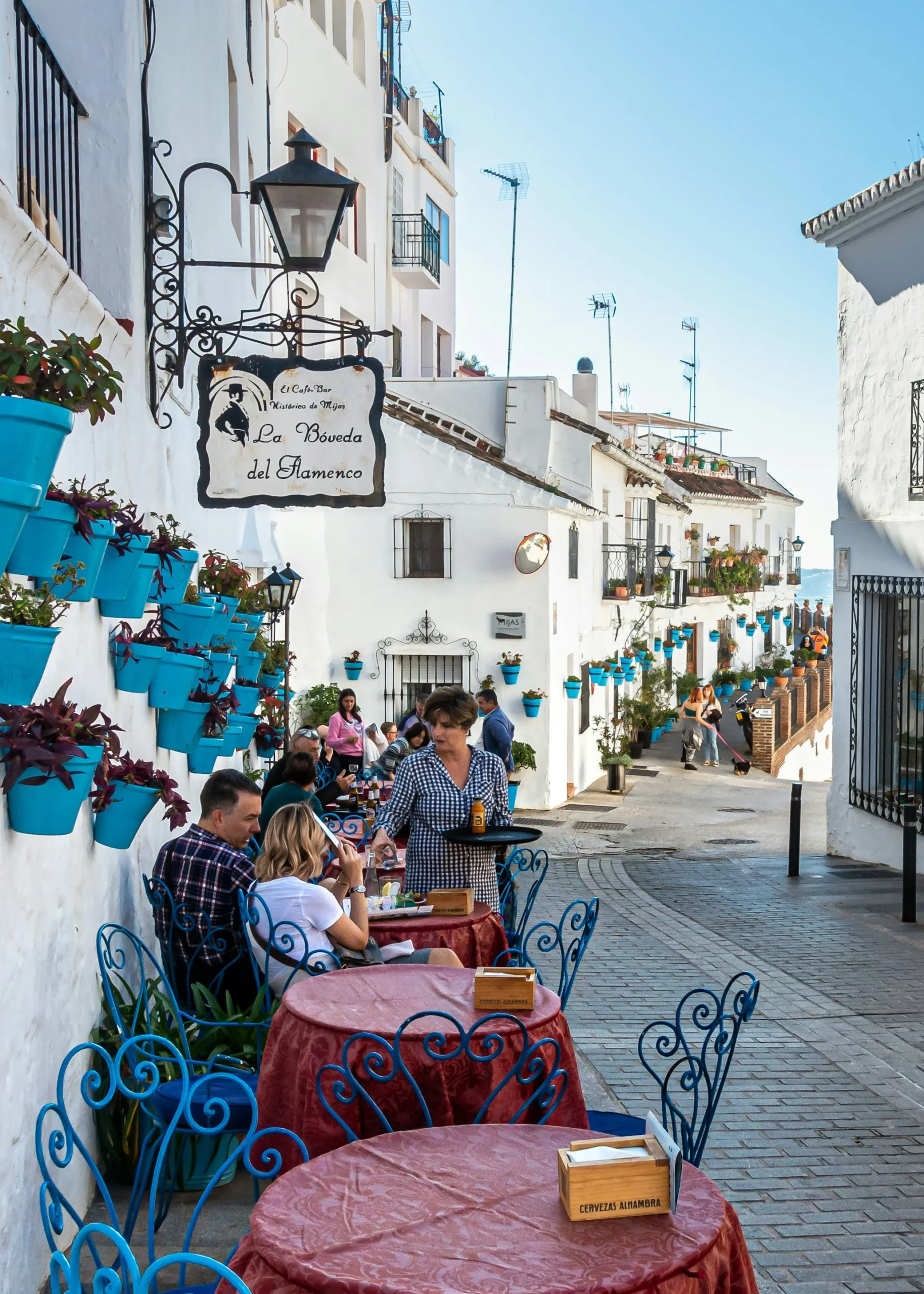 Charming cobblestone street in Mijas with white buildings, bright blue flower pots, and tourists dining at outdoor cafe tables