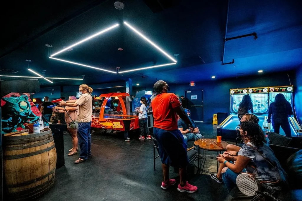 People playing arcade games, air hockey, and skeeball inside a dimly lit entertainment venue with neon overhead lighting