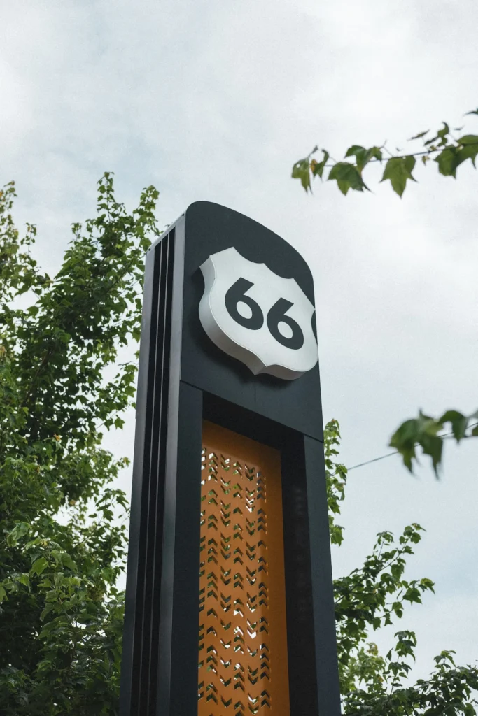 Modern black and yellow Route 66 highway marker sign surrounded by green tree leaves under a cloudy sky