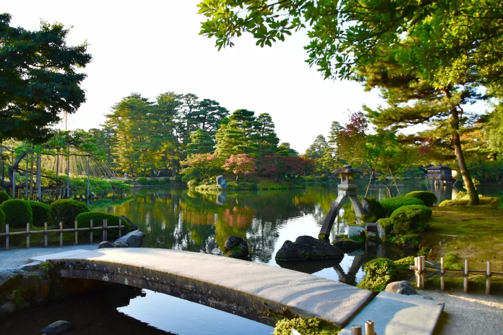 A stone bridge and Kotojitoro lantern overlooking a large reflective pond surrounded by manicured trees