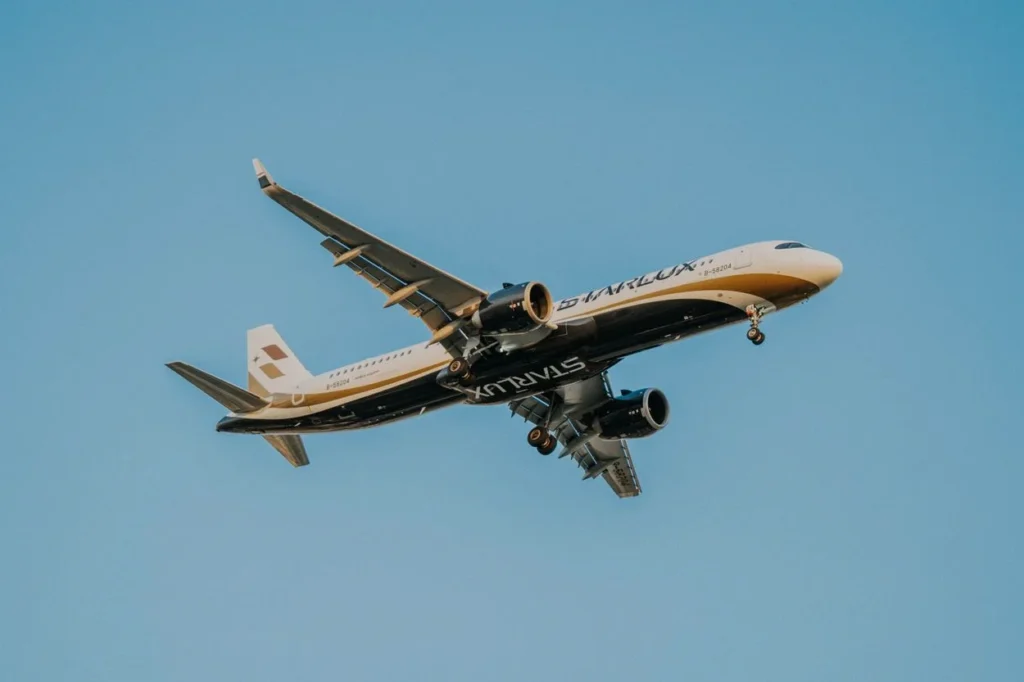 A white, gold, and dark grey Starlux Airlines Airbus A321neo passenger plane flying overhead