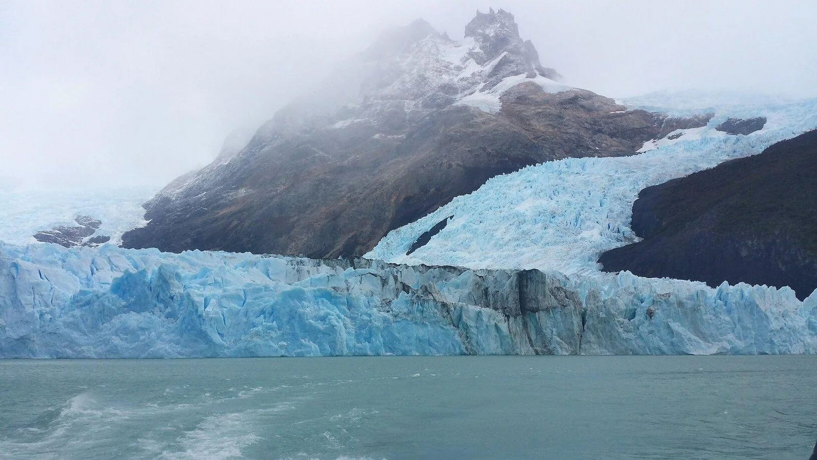Spegazzini Glacier ice wall rising from gray waters of Lake Argentino with misty Patagonian mountains in background