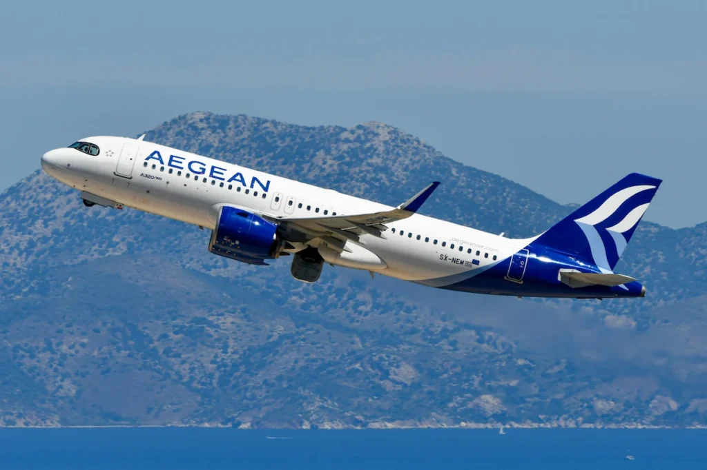 Aegean Airlines Airbus A320neo passenger jet climbing during takeoff over a bright blue sea with mountains behind