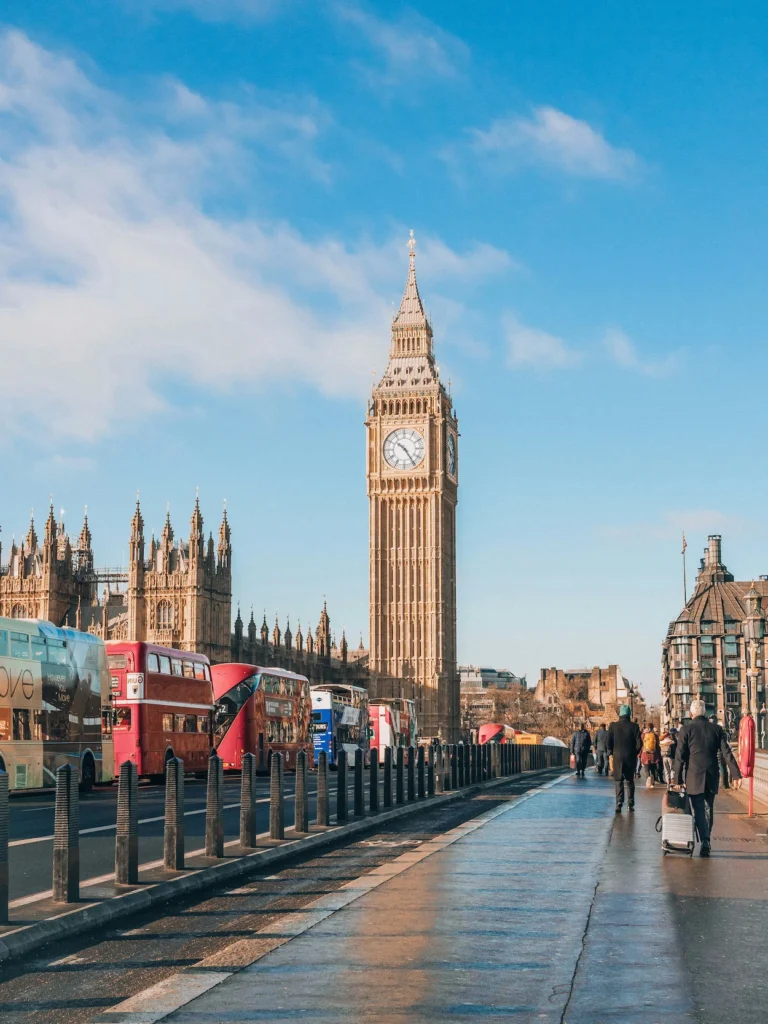 Big Ben and red double-decker buses viewed from a wet pedestrian walkway on Westminster Bridge