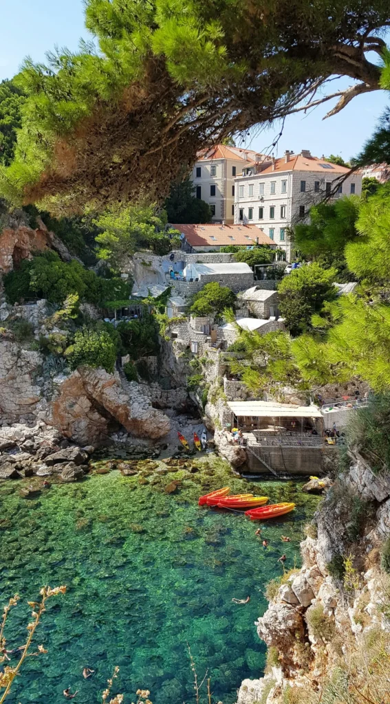 Clear turquoise cove with orange kayaks nestled below rocky cliffs and stone buildings in Dubrovnik