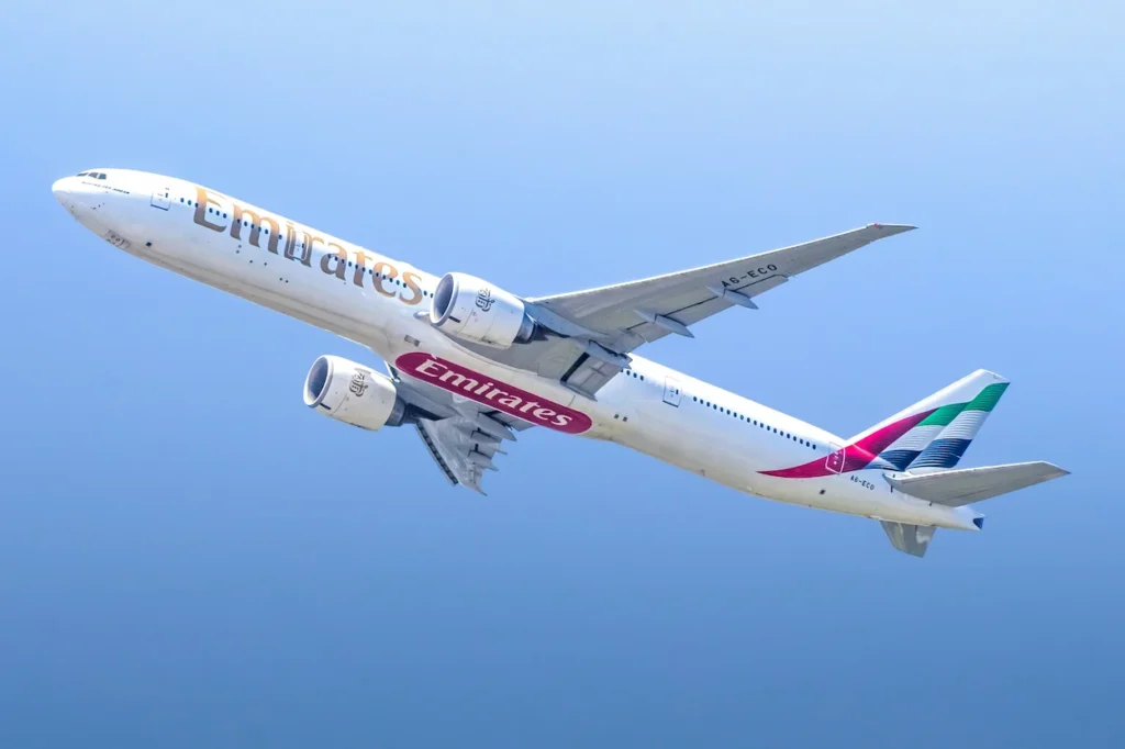 An Emirates Boeing 777-300ER commercial passenger airplane climbing steeply against a solid blue sky background after departure