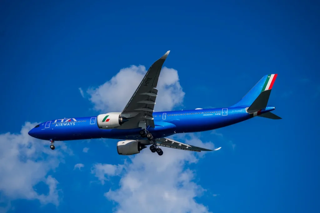 ITA Airways Airbus A330neo commercial aircraft flying overhead against a blue sky with scattered clouds