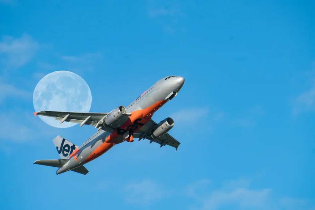 An orange and gray Jetstar Airbus A320 flying across a bright blue sky past a large moon