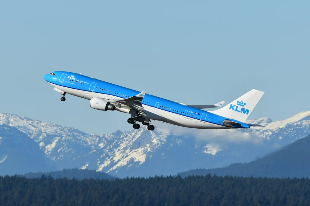 KLM Airbus A330 passenger plane climbing into the sky above a forest with snow-covered mountains in the distance