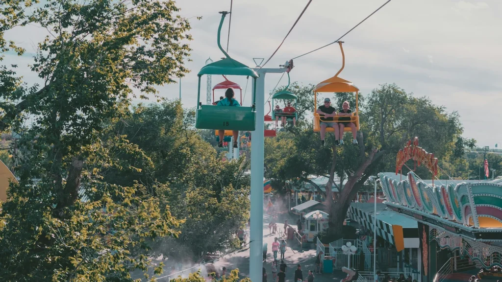 People riding colorful open-air sky ride chairs over a busy amusement park midway with green trees and carnival games