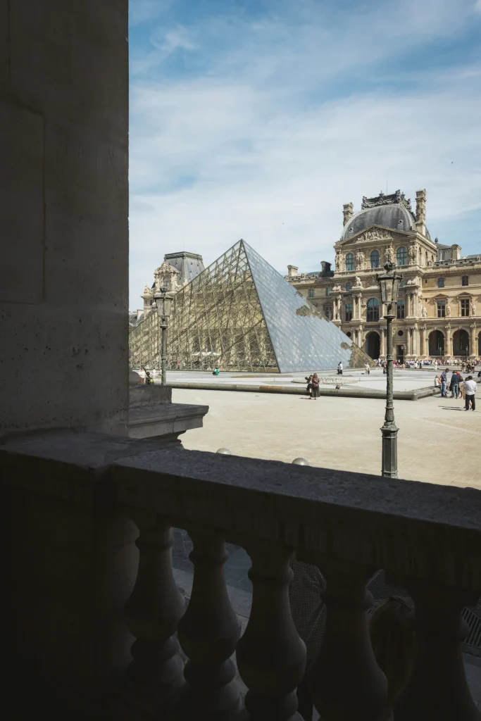 Louvre glass pyramid and classical palace facade framed by a stone balcony in the foreground
