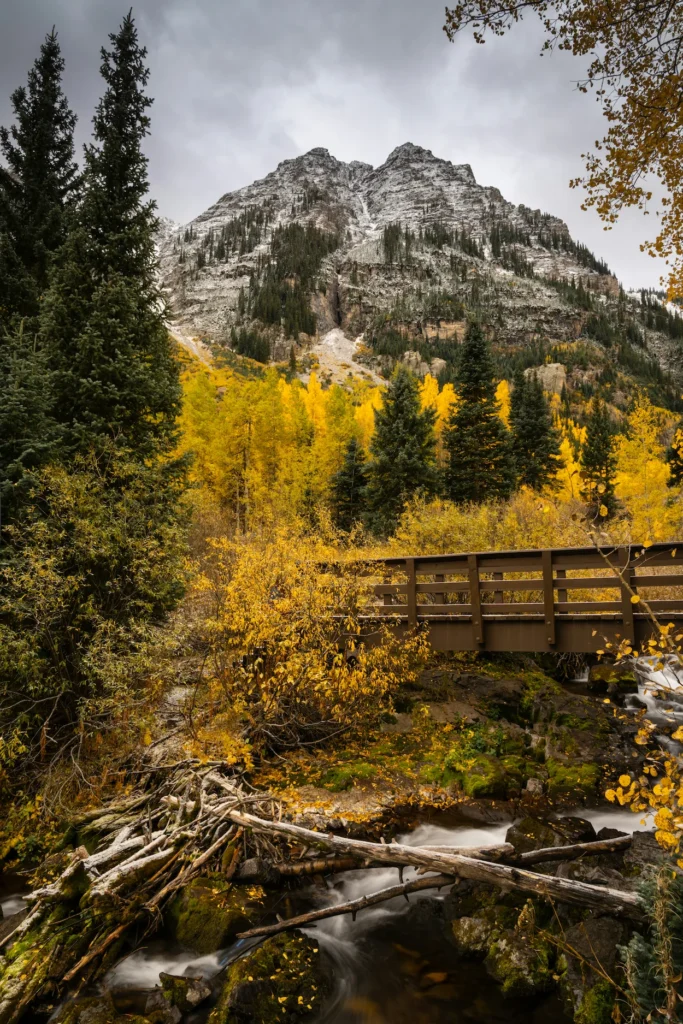 Wooden footbridge over a creek with yellow autumn trees and a snow-dusted peak at Maroon Bells in Aspen