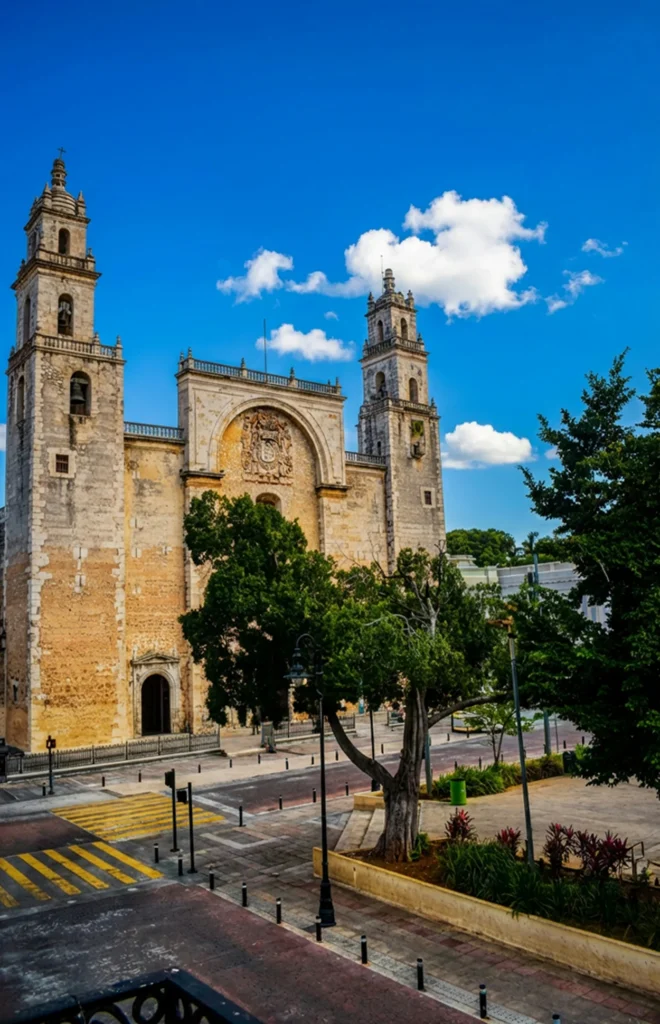 The stone facade and tall bell towers of the Mérida Cathedral overlooking a plaza street under a vibrant blue sky