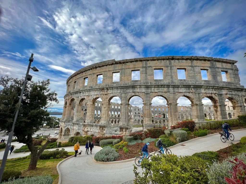 Pula Arena Roman amphitheater stone walls next to a pathway with cyclists under a blue sky