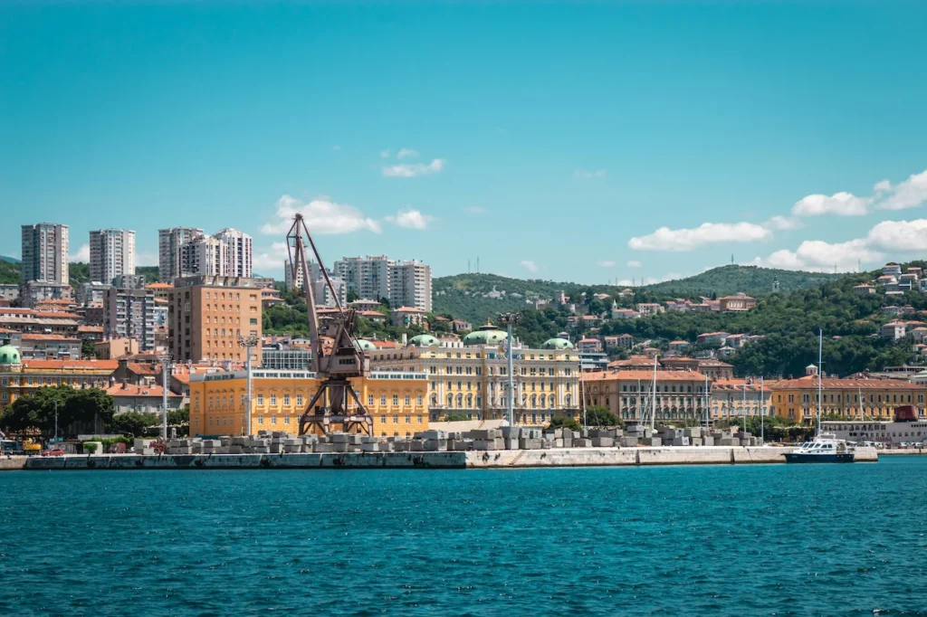 Rijeka harbor pier with a large industrial cargo crane in front of yellow coastal buildings and green hills