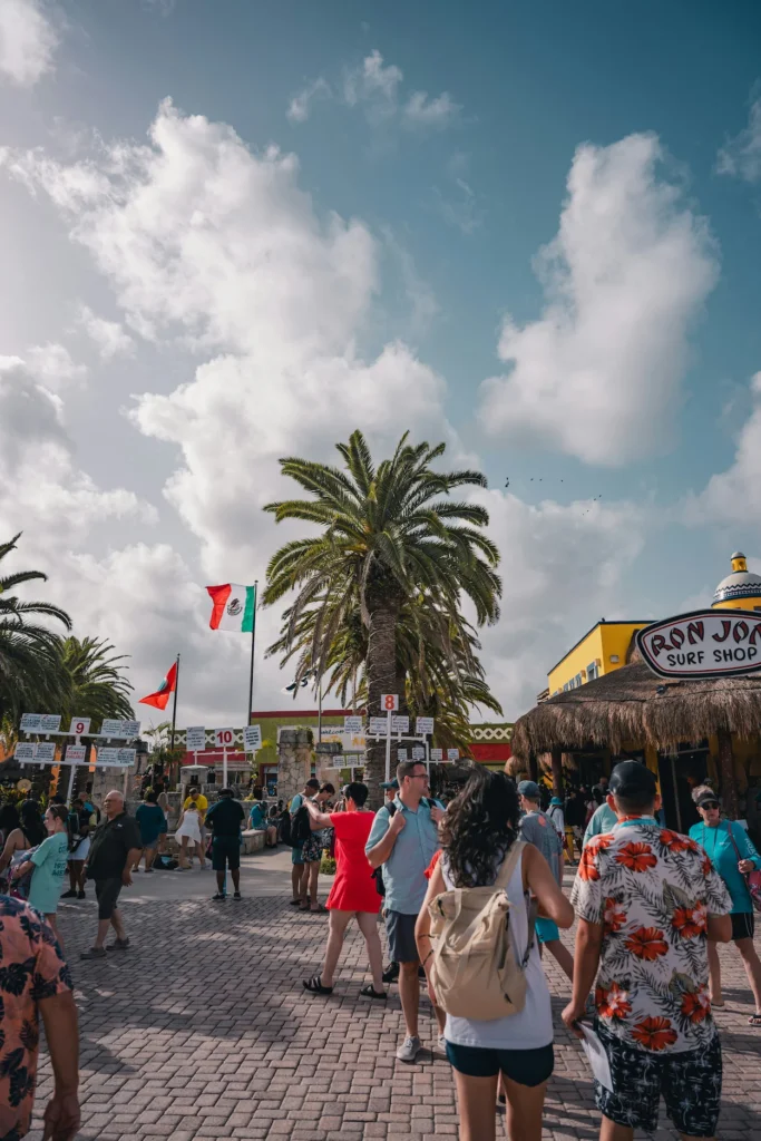 Tourists walking through a sunny Cozumel port plaza next to a Ron Jon Surf Shop under a blue sky