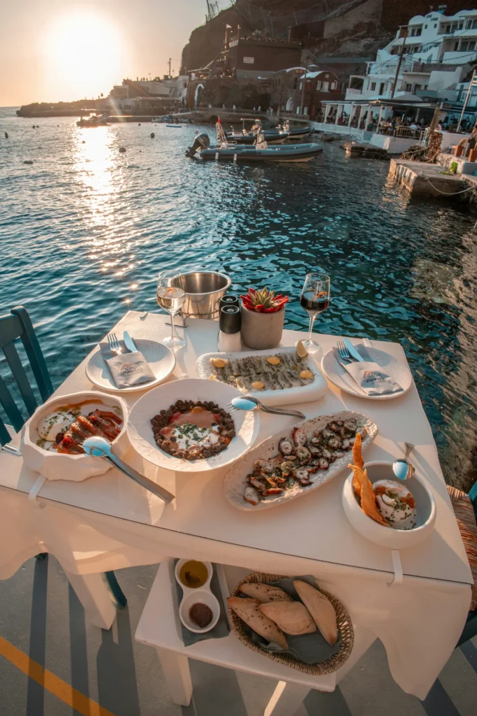 Outdoor restaurant table set with Greek seafood dishes and wine next to the sea in Santorini at sunset
