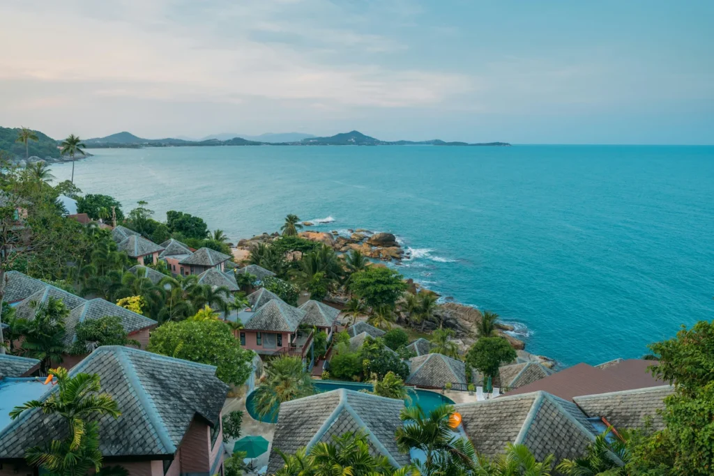 Thatch-roofed luxury villas clustered on a green hillside overlooking the ocean in Koh Samui, Thailand