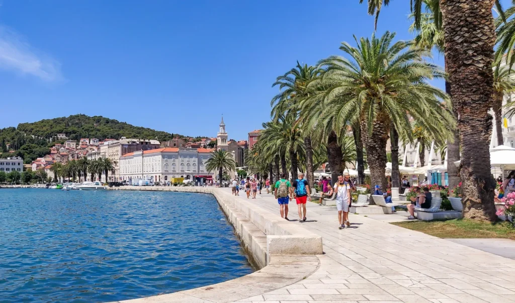 Split Riva waterfront promenade lined with tall palm trees, pedestrians, and blue Adriatic Sea water