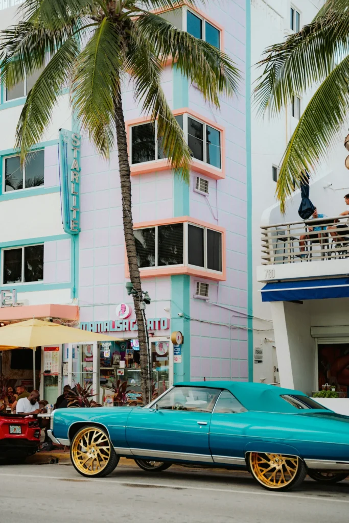 A classic teal convertible with large gold rims parked on Ocean Drive in front of the pastel pink and turquoise Starlite Hotel in Miami Beach