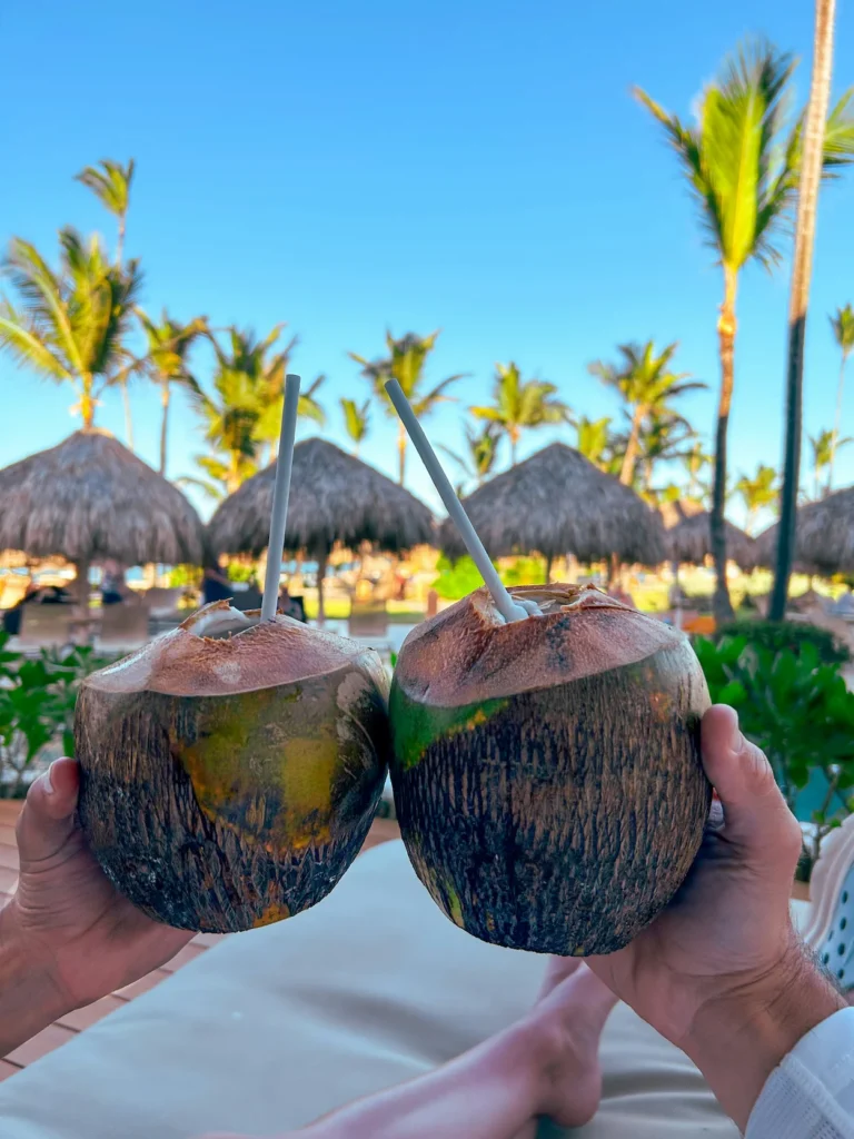 Two hands holding fresh green coconuts with straws at a tropical beach resort