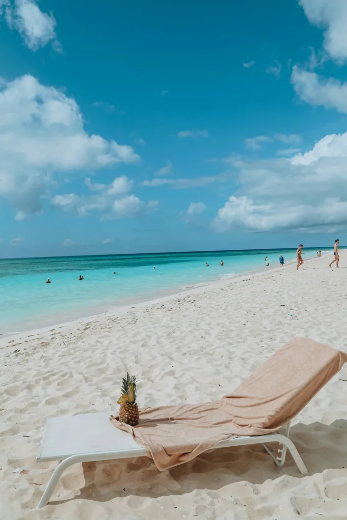 A lounge chair with a towel and a pineapple on a wide white sand beach next to turquoise ocean water