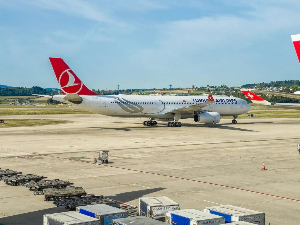 A white Turkish Airlines Airbus A330 commercial airliner parked on the apron of an airport tarmac with cargo containers in the foreground