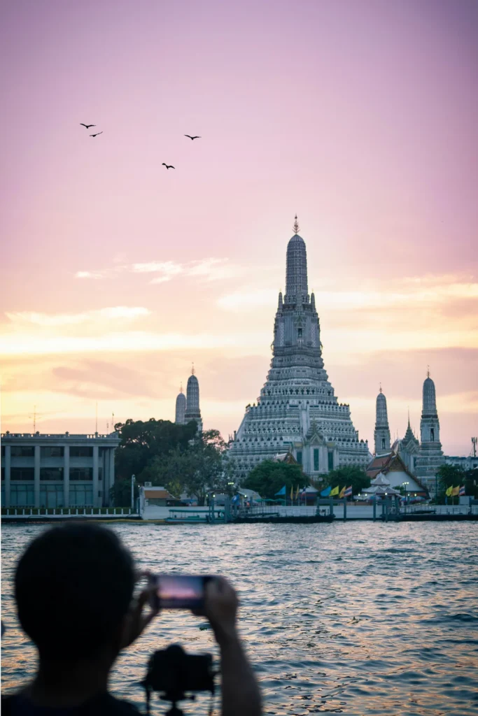 Tourist taking a photo of Wat Arun temple across the Chao Phraya River during a pink sunset in Bangkok