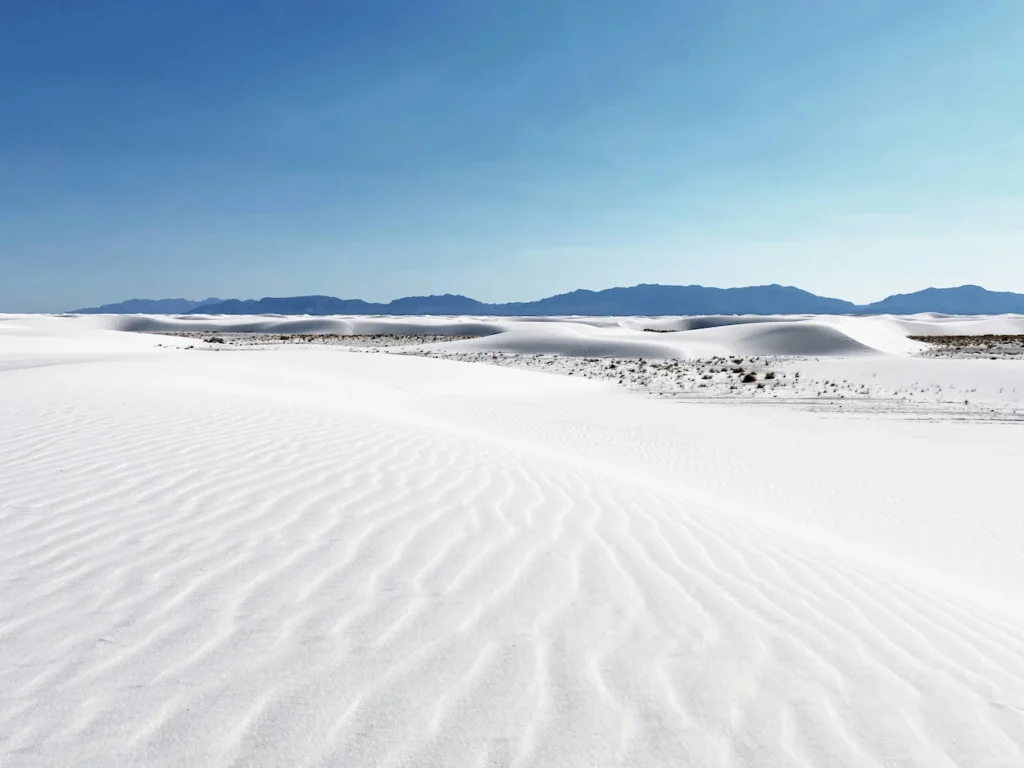 White gypsum sand dunes with textured ripples stretching toward distant blue mountains under a clear blue sky