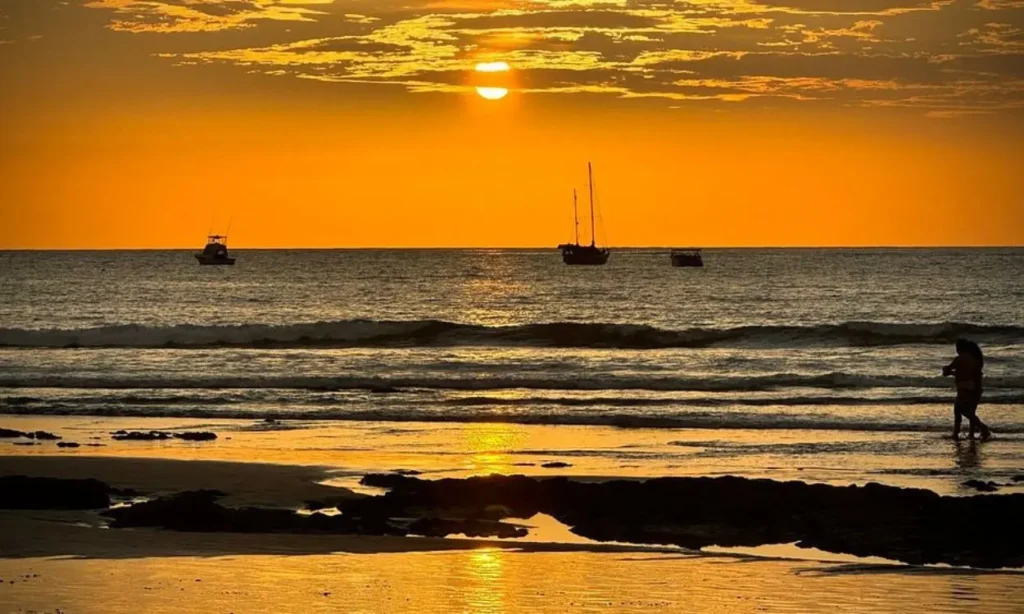 Sunset over Tamarindo Beach with boats on the water and people walking along the shore.